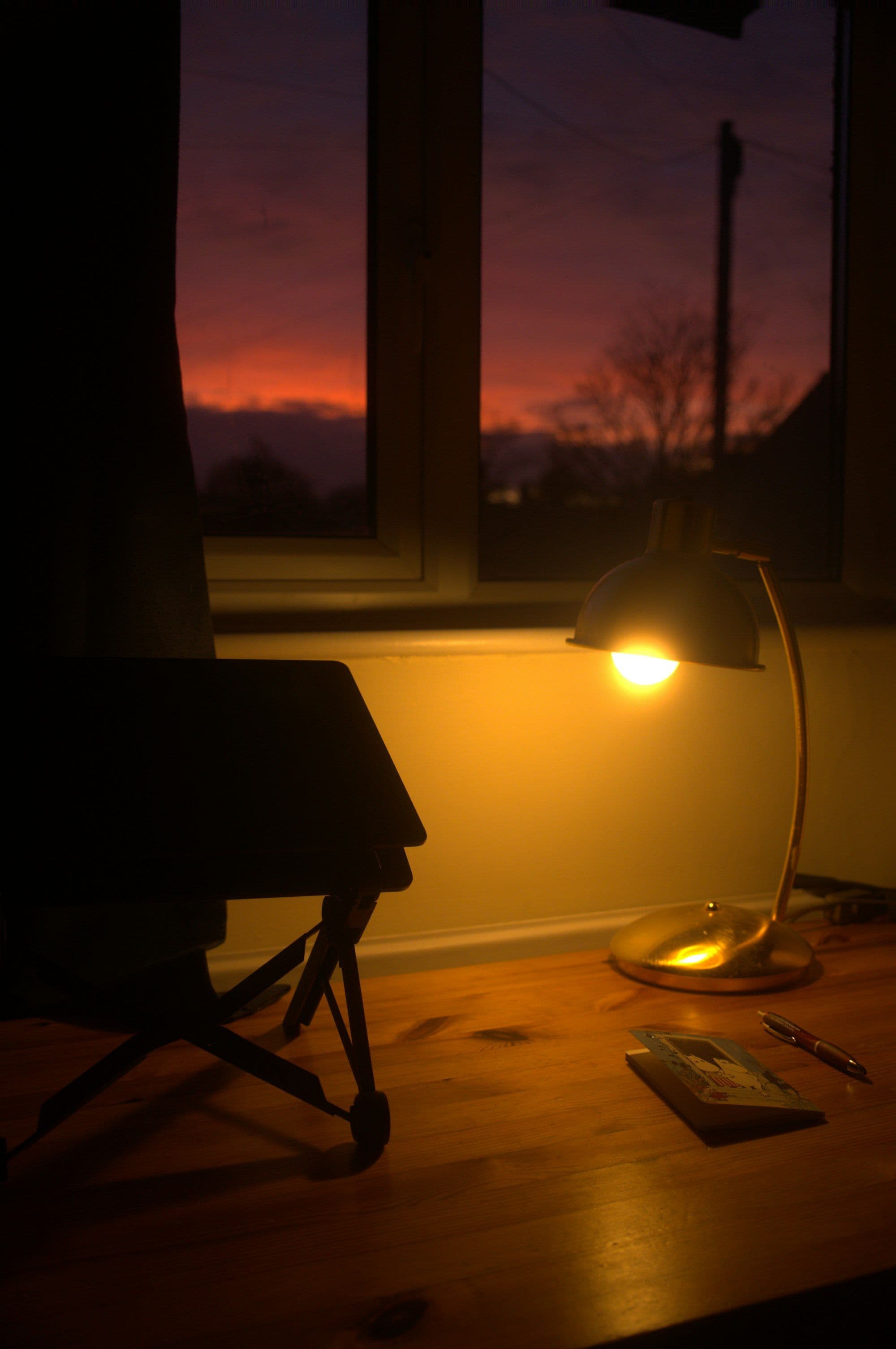 A warm desk lamp glows in a darkening room as the last light fades through the window.